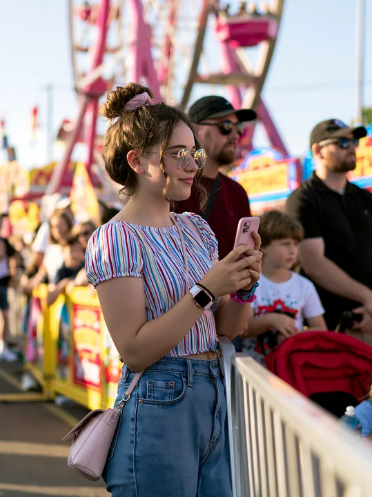 Family waits in line near a colorful ferris wheel at a sunny outdoor fair.