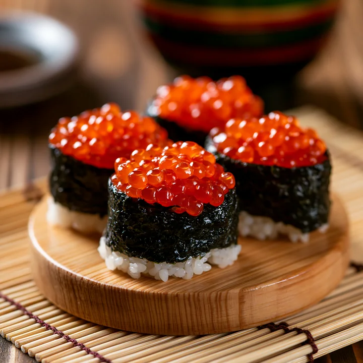 Four pieces of sushi topped with bright orange ikura salmon roe are arranged on a round wooden plate, with a bamboo mat background.