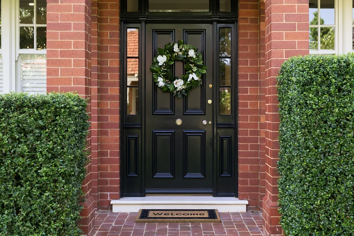 Front door with wreath and welcome doormat stands at the entry of a brick house.