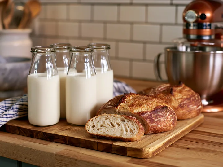 Glass milk bottles arranged beside fresh bread on a wooden cutting board in a kitchen.