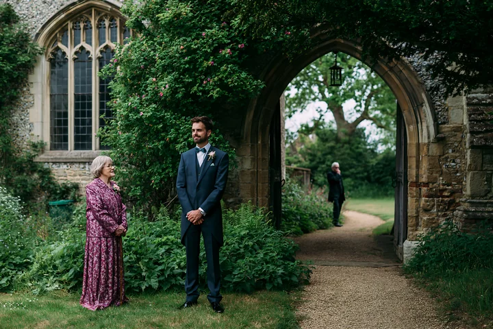 Groom waits beside an older woman beneath a stone church arch in a leafy garden.