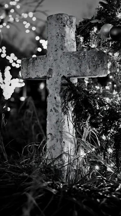 A handcrafted Christian cross stands among grass in a Christmas meadow. The scene is lit by decorative lights and surrounded by greenery, creating a festive atm