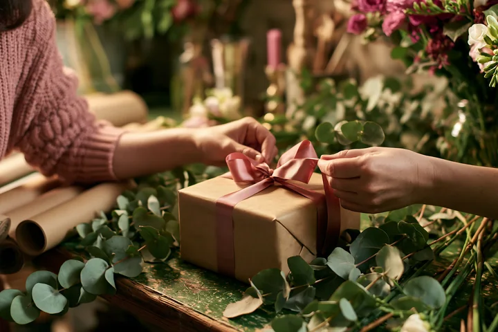 Hands are tying a ribbon around a gift box placed on a table.