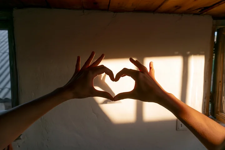 Hands create a heart shape casting a shadow on a wall in a warm room with wooden features.