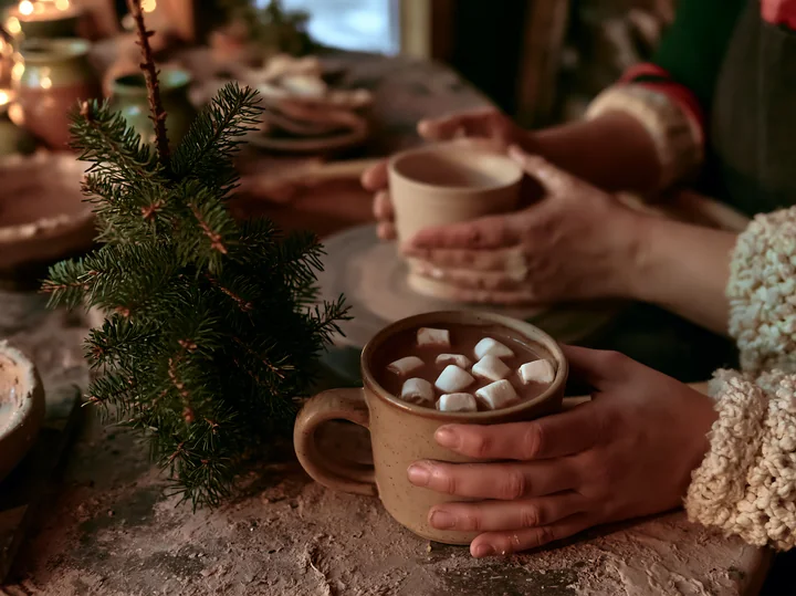 Hands hold a mug of cocoa topped with marshmallows beside a small evergreen tree at a warmly lit table.
