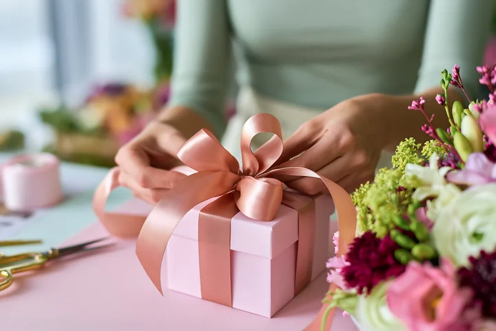 Hands tie a pink ribbon around a gift box beside a colorful flower arrangement.