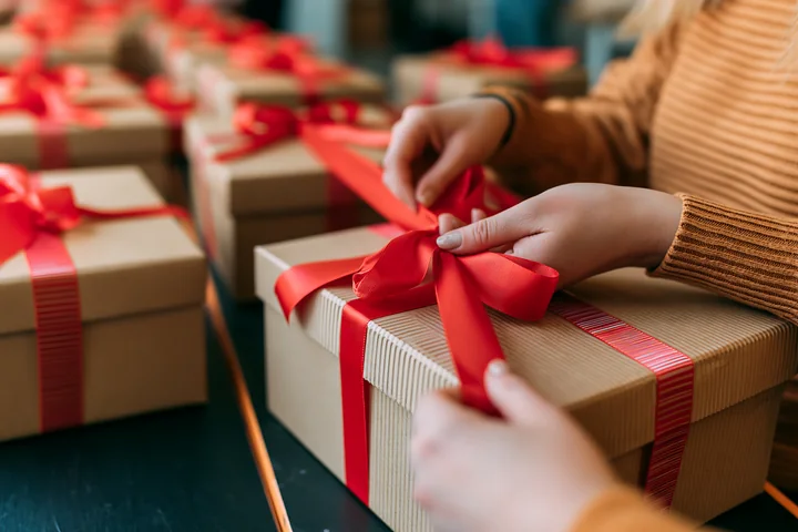 Hands tie a red ribbon around a gift box in a holiday setting filled with wrapped presents.