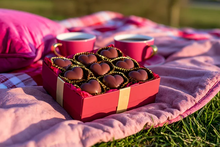 Heart-shaped chocolates in a red gift box rest on a pink blanket beside two cups outdoors.