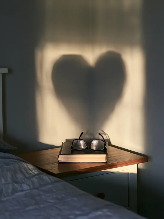 Heart-shaped shadow from window light falls on a bedroom wall behind eyeglasses and a closed book on a bedside table.