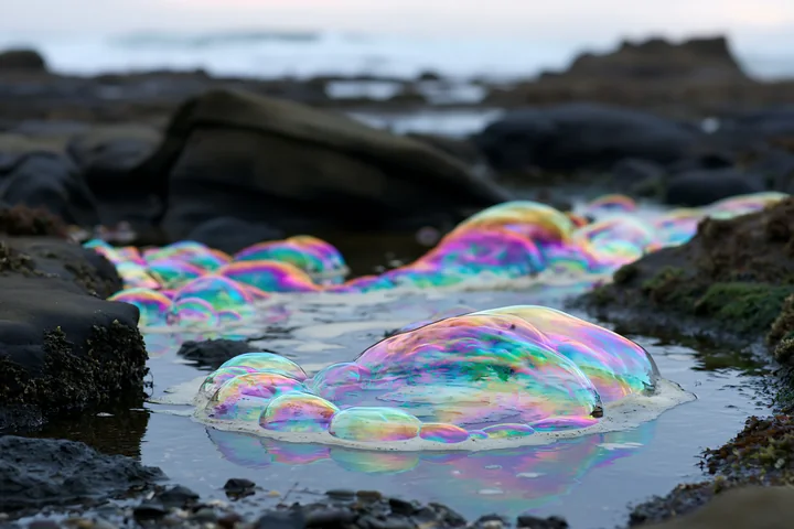 Iridescent bubble foam spiral collects along a rocky shore at low tide.