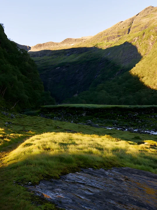 Sunbeams light up the meadow in Jotunheimen valley. The scene shows soft green grass and rocky terrain under a clear sky. The mountains create a dramatic backdr