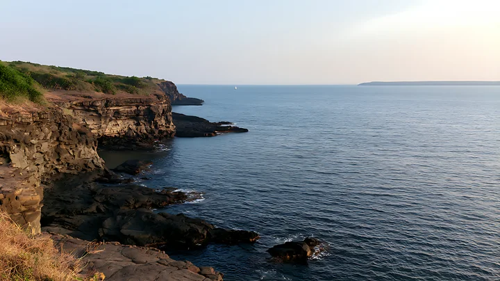 Layered coastal cliffs meet calm blue ocean under a bright open sky, with a small sailboat offshore.
