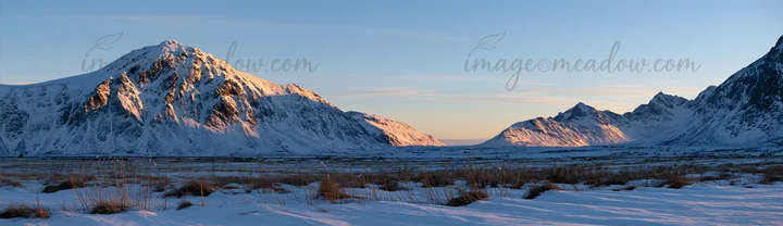 The scene shows snow-covered meadows in Lofoten, surrounded by mountains under a clear blue sky during winter. The landscape highlights the rugged terrain and d