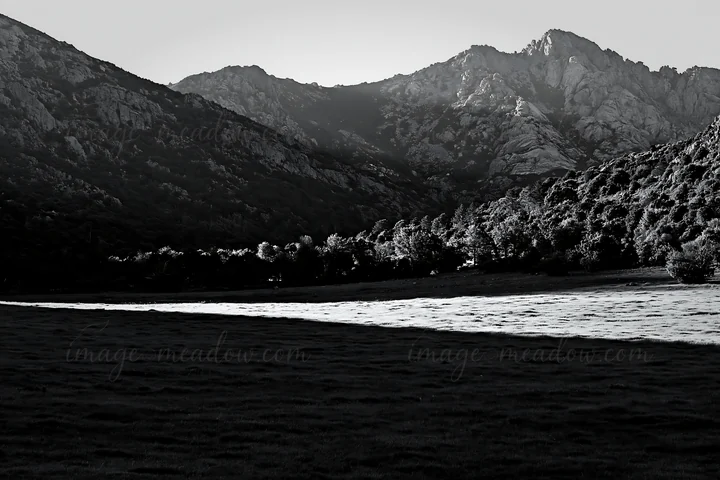 A meadow surrounded by mountains during the day. Sunrays shine down on the grassy area, creating a bright contrast against the dark mountains in the background.