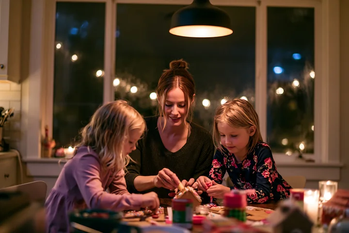 Mother and two daughters assemble and decorate a gingerbread house at a warmly lit dining table.