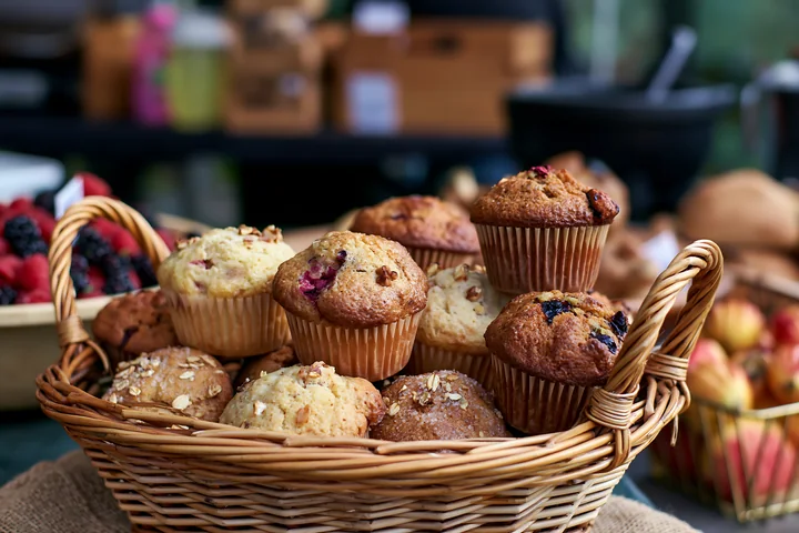 Muffins are arranged in a basket at a bakery with other baked goods seen in the background.