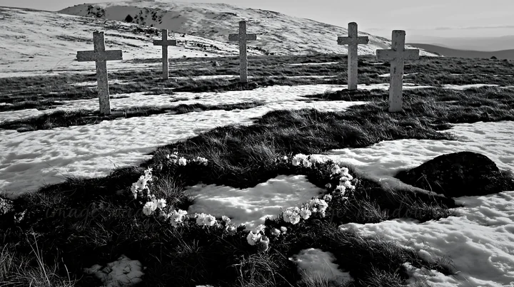 Five Christian crosses stand in a snowy meadow. A heart-shaped arrangement of flowers is in front of the crosses, set against a backdrop of mountains. The scene