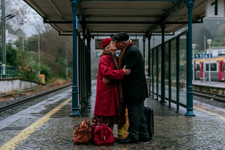 Older couple kissing beside luggage on a rainy train station platform.