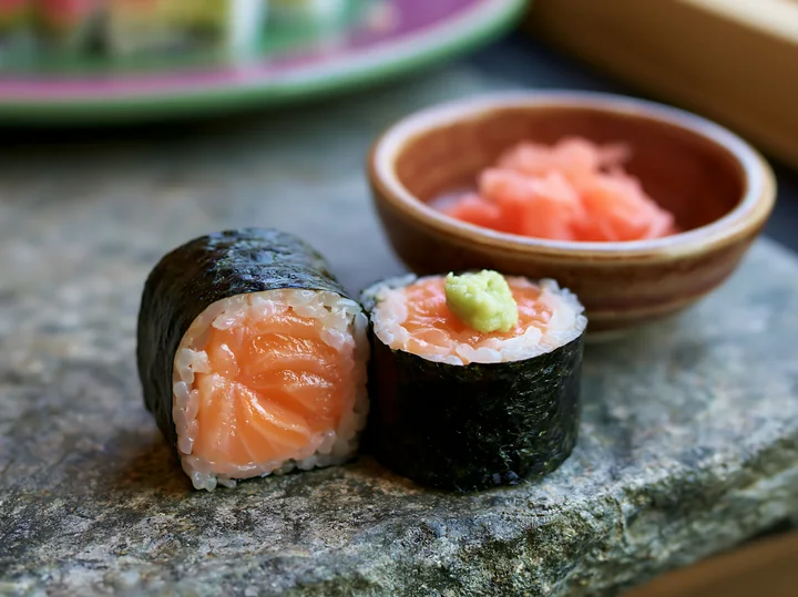 One salmon maki piece with wasabi sits on a stone surface next to a bowl of pickled ginger.