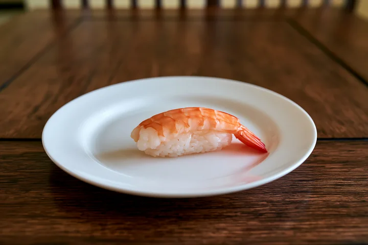 One shrimp sushi piece on a white plate sits on a wooden table.