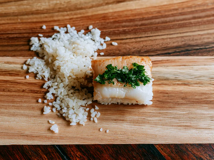One sushi piece with fresh herb garnish sits next to a small mound of rice on a wooden board.