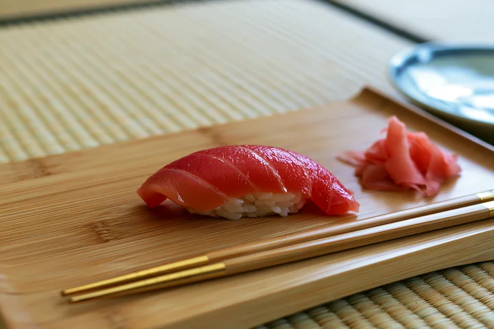 One sushi piece with pickled ginger rests on a bamboo plate beside chopsticks.