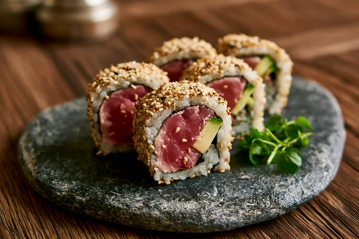 One tuna roll slice with sesame coating is placed on a dark stone plate alongside three other sushi pieces.