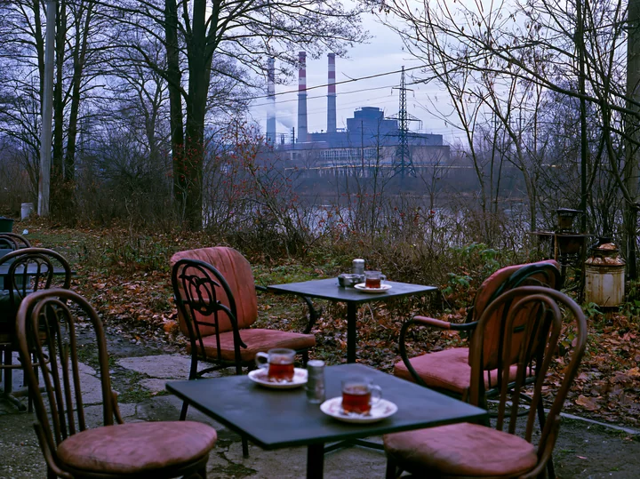 Outdoor seating shows two tables with drinks, while Soviet factory smokestacks rise in the background.