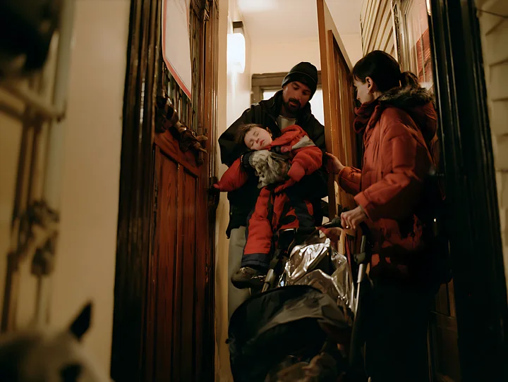 Parents carry a sleeping child in a stroller through a warmly lit interior hallway.