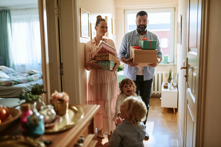 Parents carry decorated gift boxes into a bright home while three children greet them.