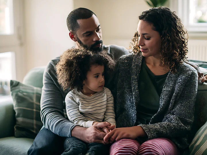 Parents comfort their young child together while seated on a sofa in a warmly lit living room.