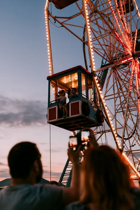 Parents photographing children on rides as they enjoy a ferris wheel at dusk.