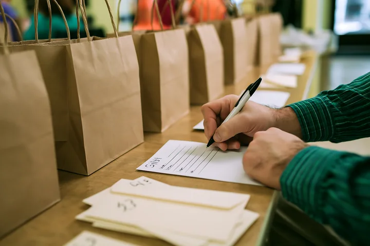 Person writes a gift list with a pen at a desk beside paper shopping bags.