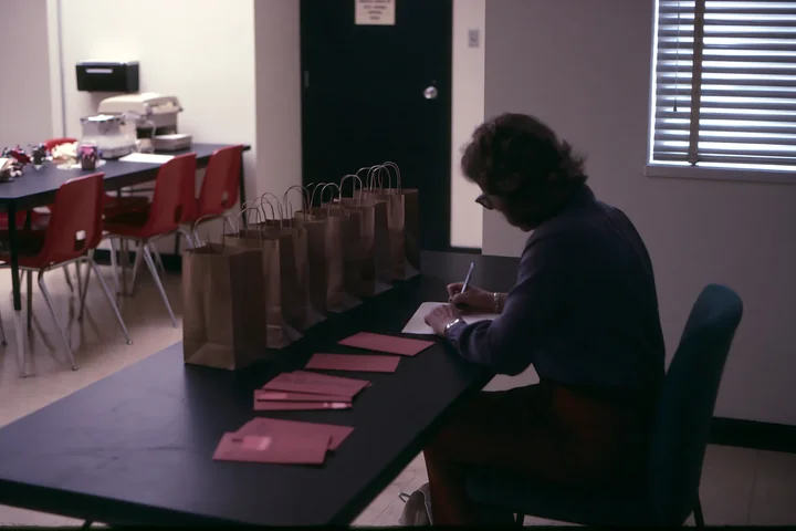 Person writes on a paper gift list at a desk beside gift bags and envelopes.