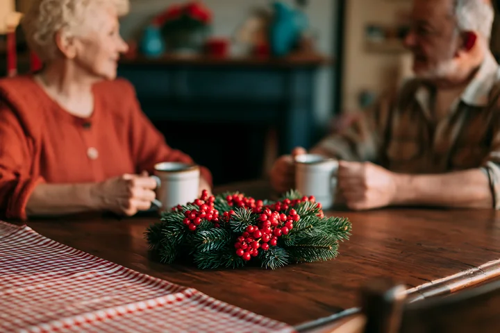 Pine wreath with red berries rests on a wooden dining table while an older couple holds mugs in the background.