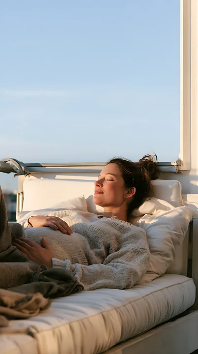 Pregnant woman rests with her hands on her belly in bed beside a sunlit window.