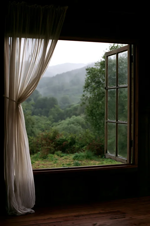 Rain drips on a cabin window while the view shows a green mountain landscape under a gray sky.