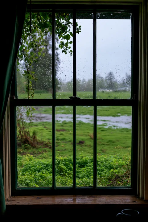Rain on small cottage window overlooking field shows drops sliding down the glass.