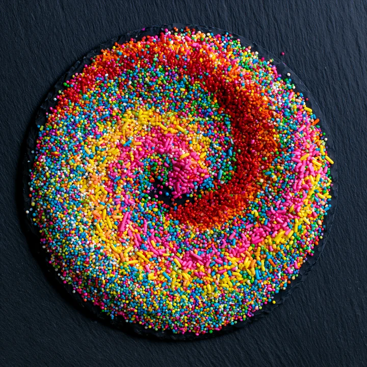 Rainbow sprinkles arranged in a swirl create a vibrant pattern on a black surface, showcasing many colors and sizes of sprinkles, ideal for dessert decoration o