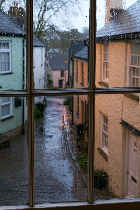 Raindrops are seen on a window, framing a view of a quaint cottage exterior.
