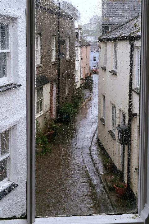 Rainy cottage exterior seen through wet glass shows a narrow street lined with buildings.