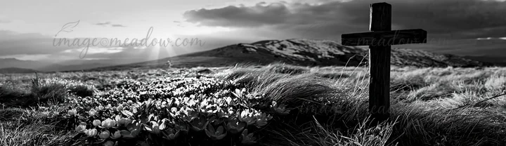 A rustic Christian cross stands in a winter meadow with snow-covered hills in the background. The sun sets behind the mountains, creating a dramatic sky filled