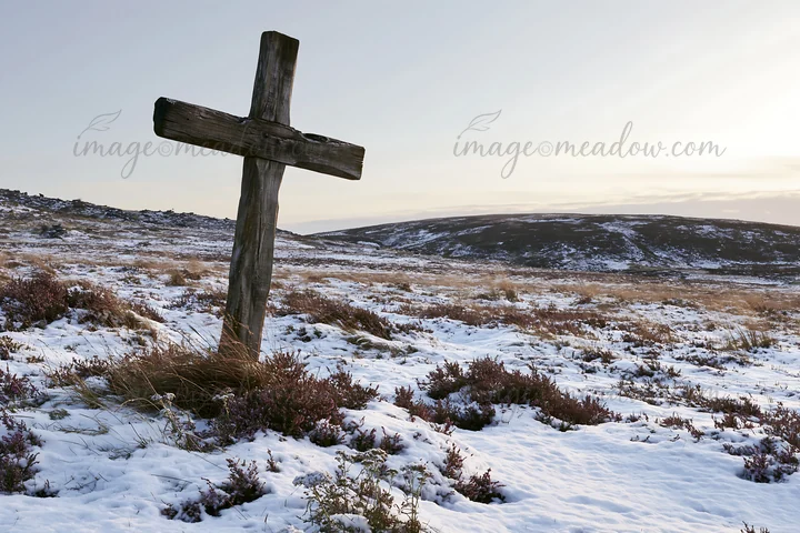 A wooden cross stands in a snow-covered meadow during winter. The landscape features dry grass and distant hills under a clear sky. This scene captures a quiet