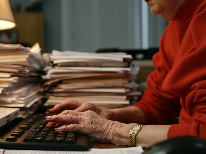 Senior woman typing with shaky hands at a cluttered desk surrounded by piles of paperwork.