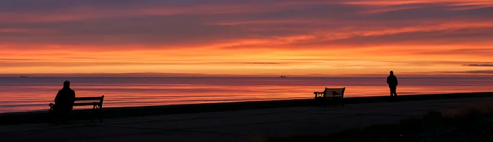 Silhouettes sit and stand beside seaside benches during a vivid orange sunset.