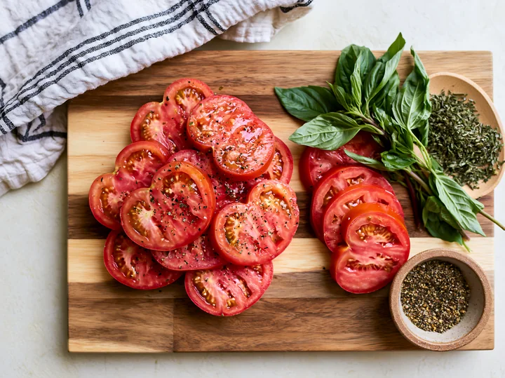 Sliced tomatoes, basil, and seasoning arranged on a wooden cutting board for cooking preparation.