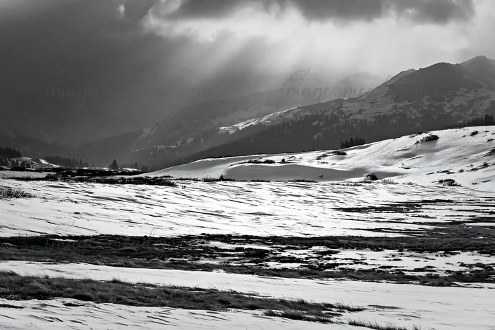 A snow-covered meadow stretches across the landscape with mountains in the background. Dark clouds loom above, casting shadows on the white ground, creating a u