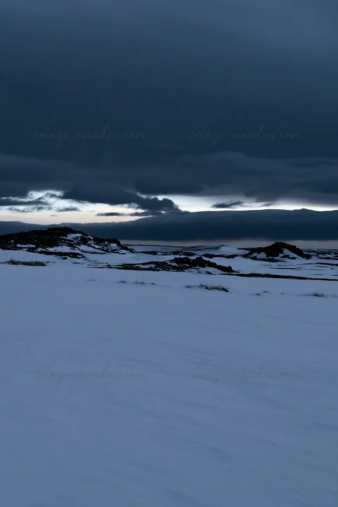 A snow-covered meadow stretches across a winter landscape. Dark clouds fill the sky, creating a dramatic scene above the white terrain. The location shows littl