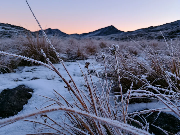 Snow covers the ground in a highland meadow. Frost clings to the grass as the sun rises, casting a soft light on the mountain range in the distance. Nature show