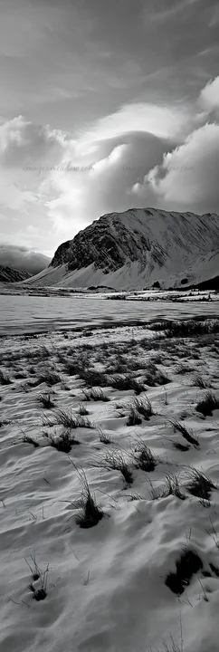 A snowy meadow borders a lake with mountains in the distance. Cloudy sky above adds depth to the scene. Visible grass peeks through the snow, creating contrast